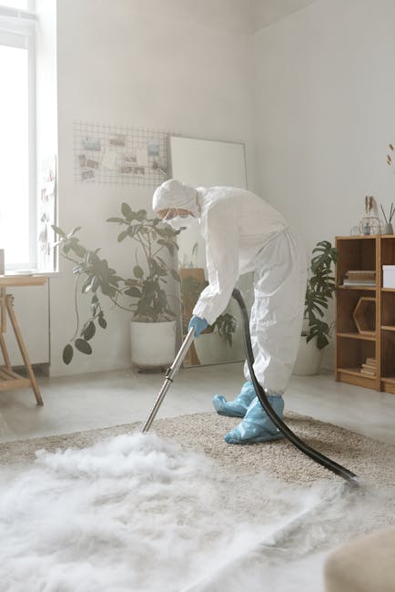 A professional cleaner from Brent Carpet Cleaners is wearing a white protective suit, gloves, and blue boot covers while performing deep cleaning on a beige carpet in a bright living room. The cleaner