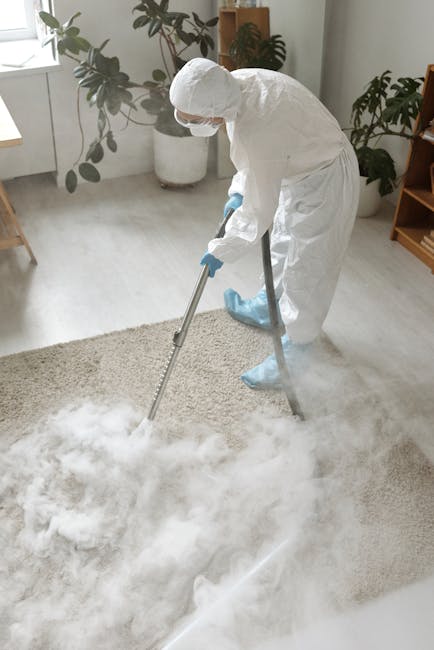 A professional cleaner from Brent Carpet Cleaners is wearing a white protective suit, gloves, and blue boot covers while performing deep cleaning on a beige carpet in a bright living room. The cleaner