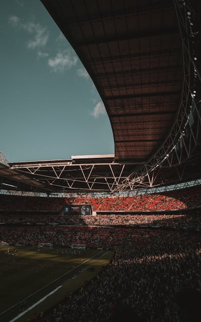 A wide view of Wembley Stadium's interior showing the football pitch with green grass and white goalposts, surrounded by red stadium seating. The roof structure is partially visible overhead with supp